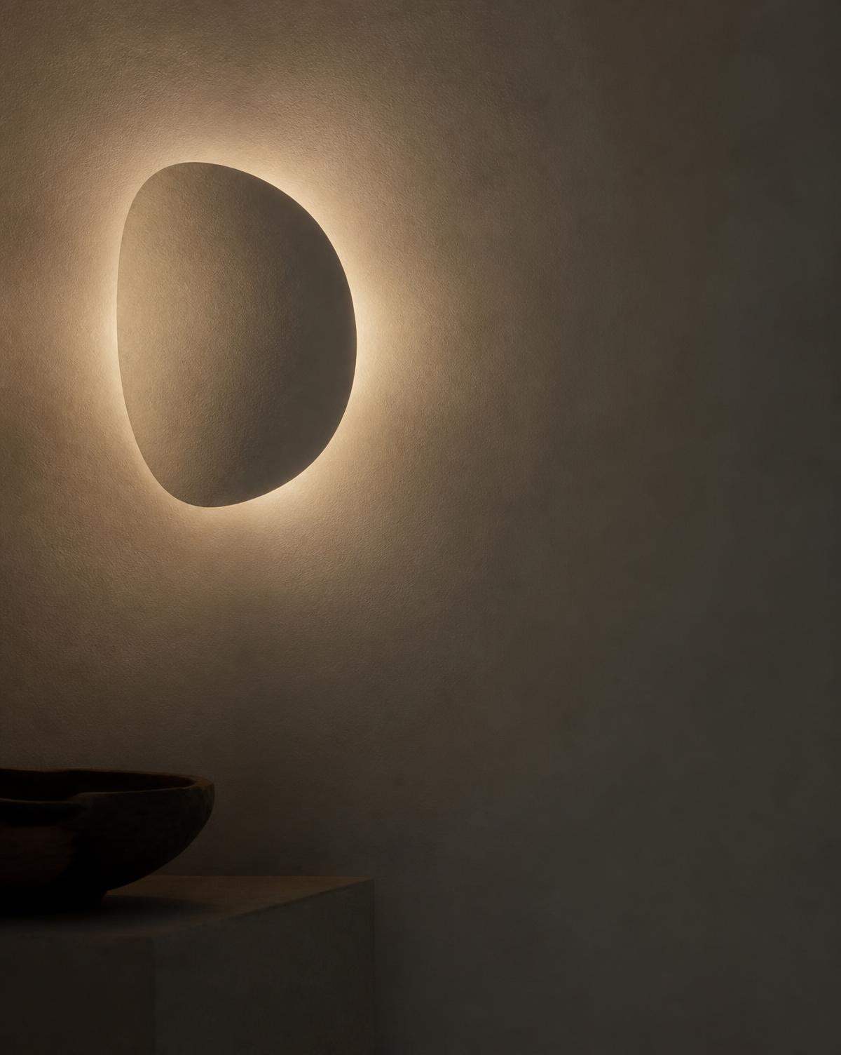 Sculptural off-white wall lamp casting a warm halo across a dark plaster wall